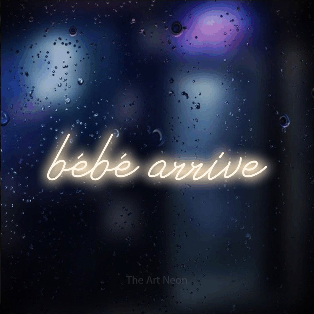 “Bébé arrive” LED neon sign in warm white, with cursive lettering, shown against glass with blue and purple reflections.