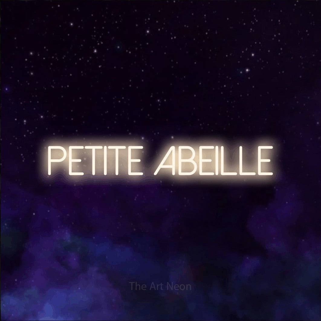 “Petite abeille” LED neon sign in warm white, with thin sans-serif lettering, shown against a purple galaxy backdrop.