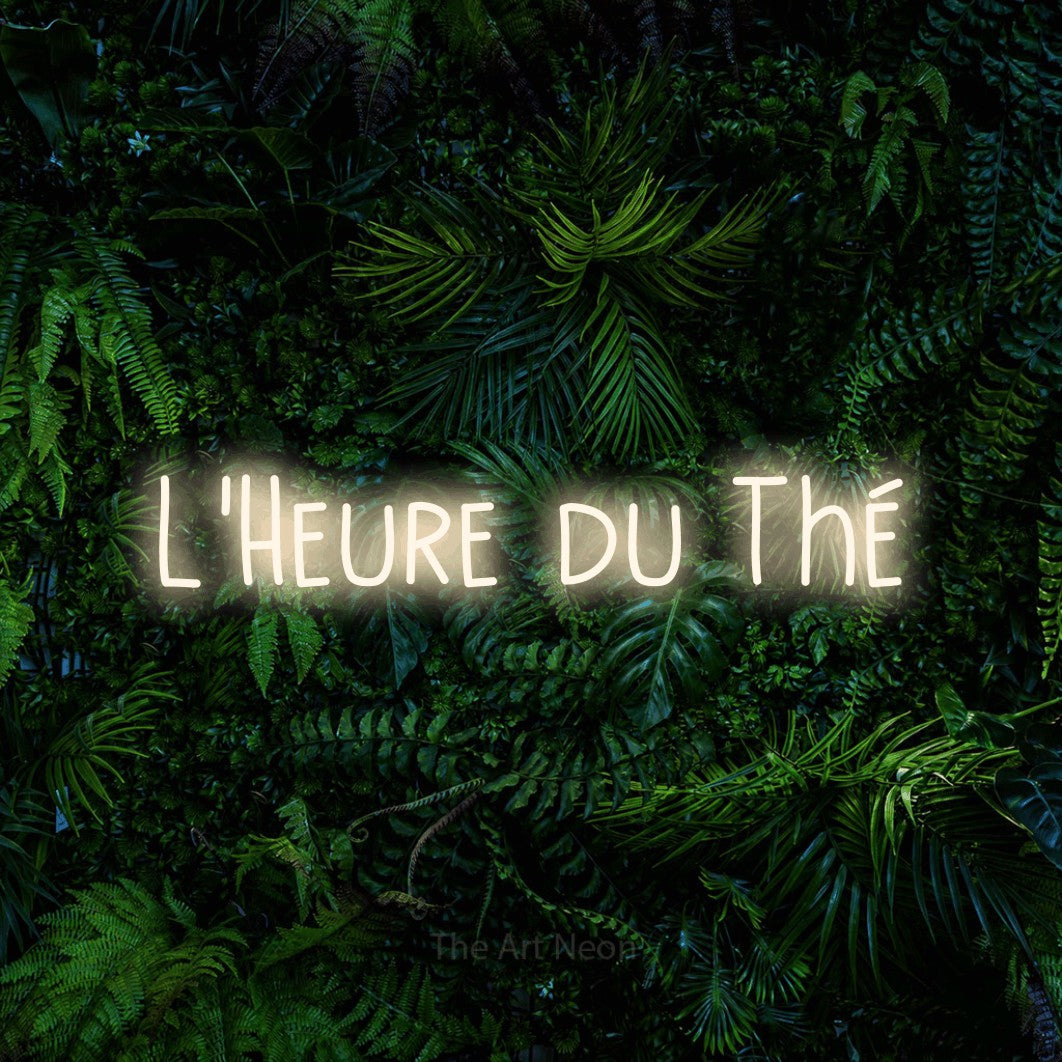 “L'heure du thé” LED neon sign in warm white, with narrow hand-drawn lettering, shown against dense tropical foliage.