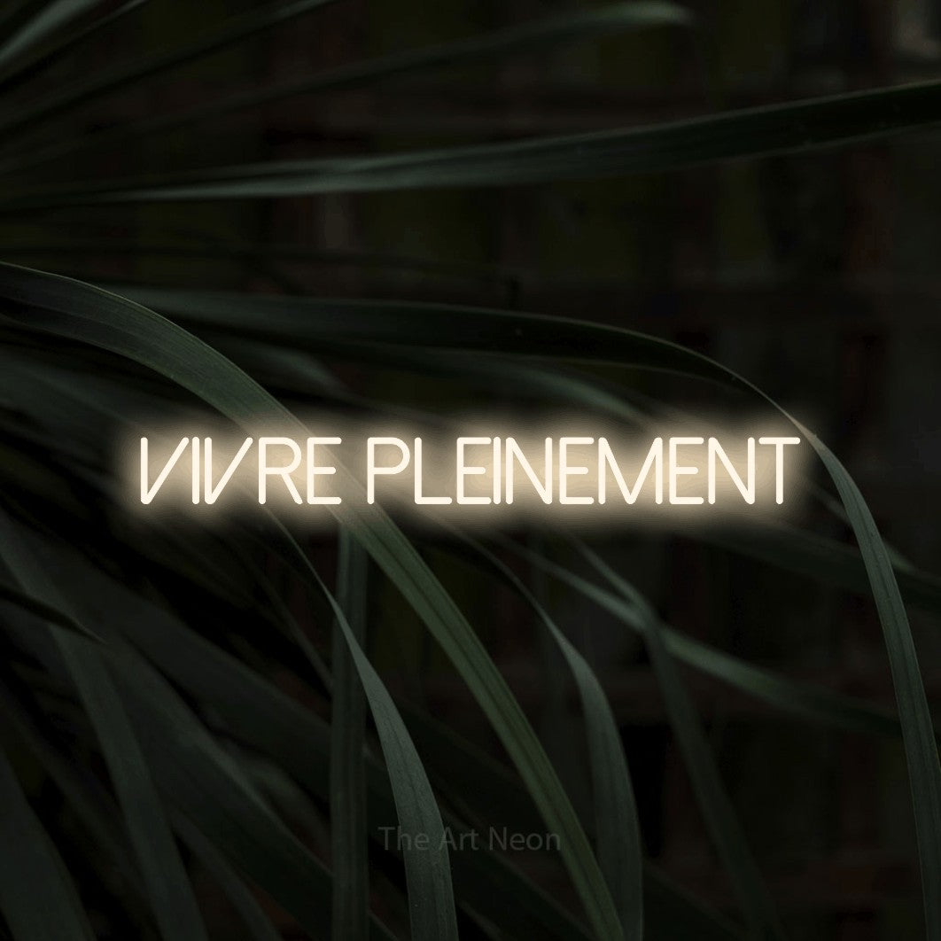 “Vivre pleinement” LED neon sign in warm white, with thin sans-serif lettering, shown against a close-up of dark foliage.