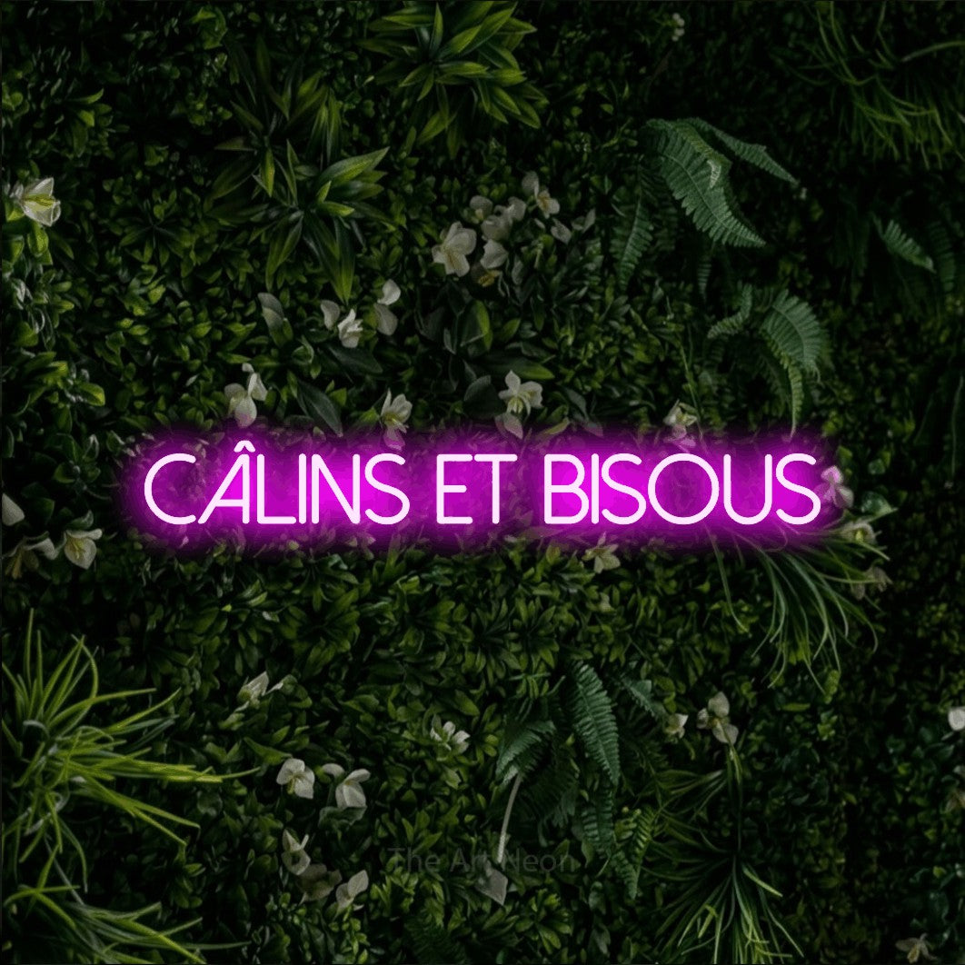 “Câlins et bisous” LED neon sign in pink, with thin sans-serif lettering, shown against a dense green plant wall.
