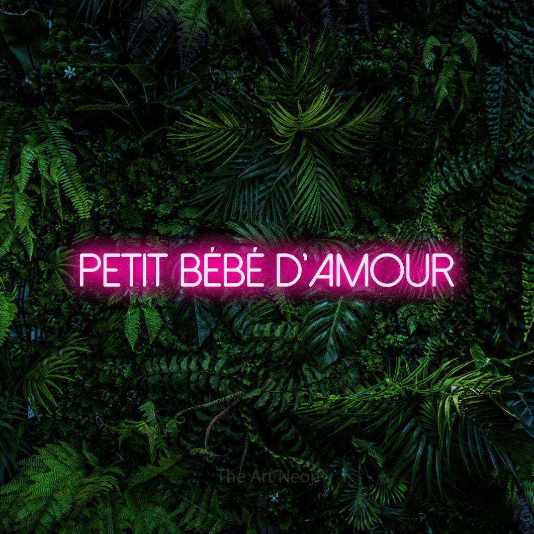 “Petit bébé d'amour” LED neon sign in hot pink, with thin sans-serif lettering, shown against dense tropical foliage.