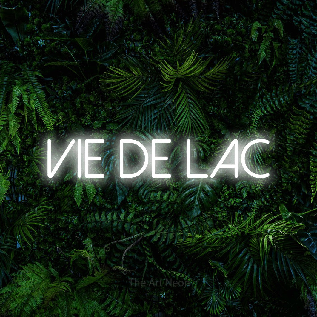 “Vie de lac” LED neon sign in cool white, with thin sans-serif lettering, shown against dense tropical foliage.