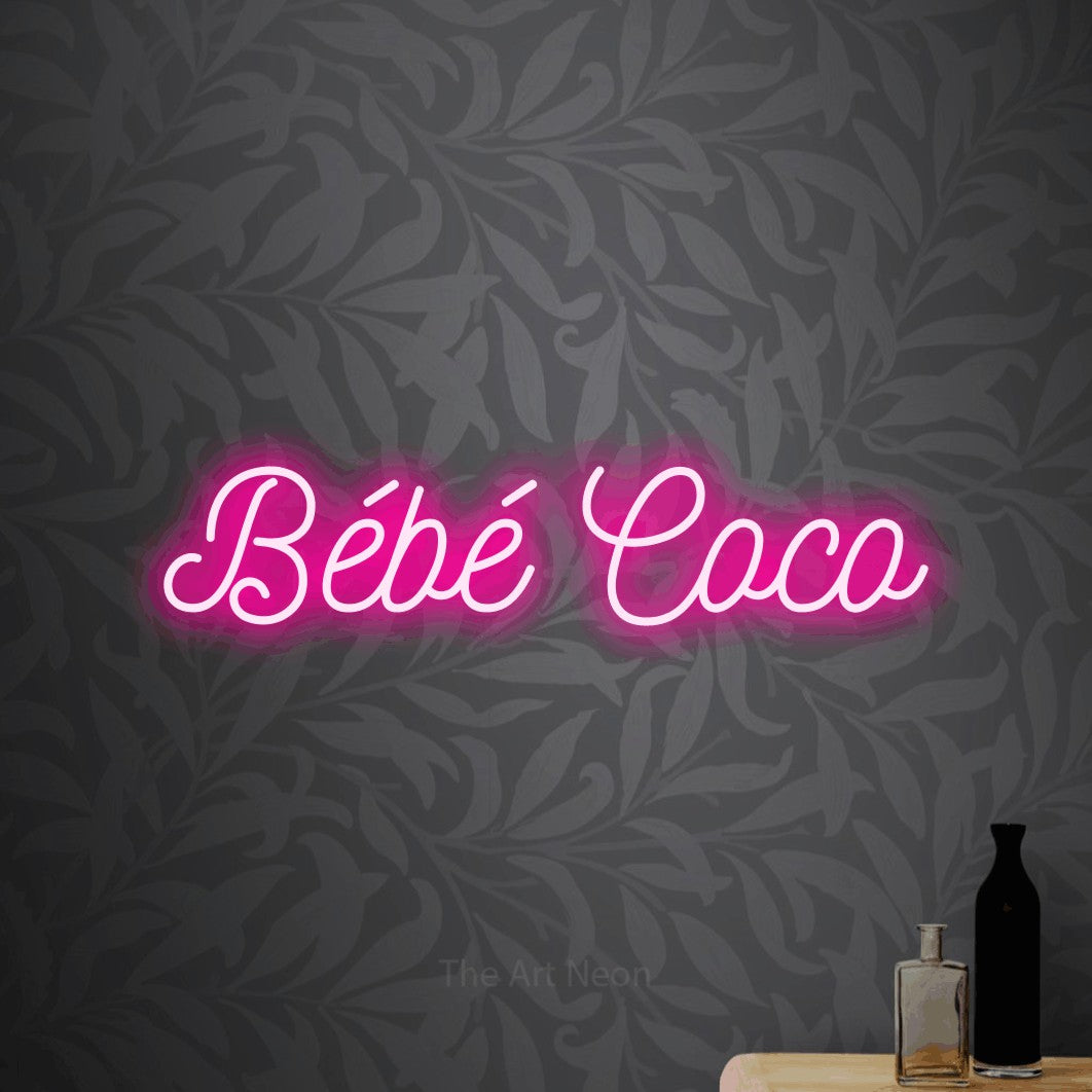 Décoration murale en néon LED « Bébé coco » en rose vif, en écriture cursive ornée, installée sur un papier peint botanique gris, pour une fête prénatale.