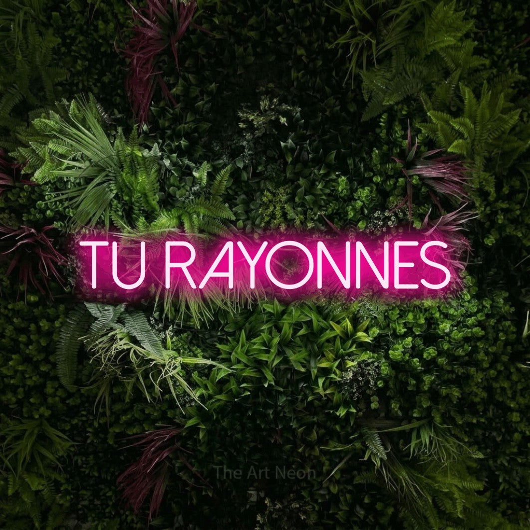 Enseigne néon LED « Tu rayonnes » en rose vif, en lettres fines sans empattement, présentée sur un mur végétal aux fleurs roses, pour un salon de beauté.
