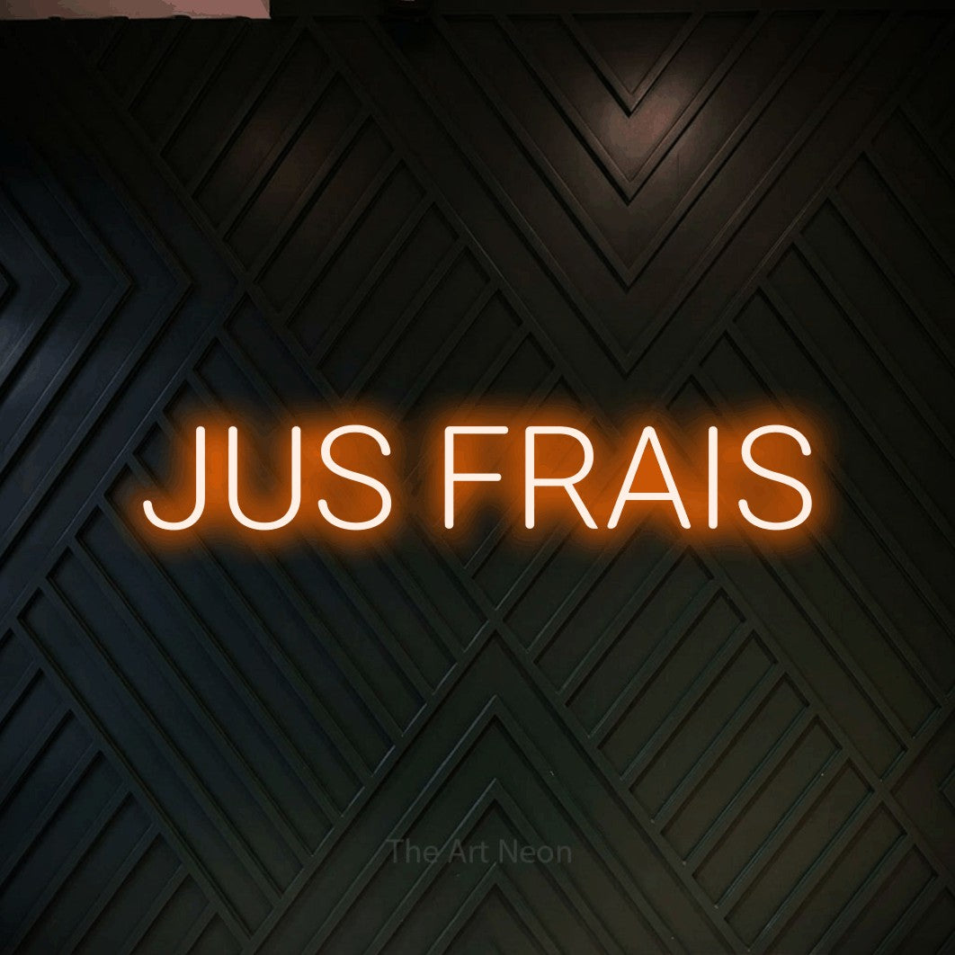 “Jus frais” LED neon sign in orange, with rounded sans-serif lettering, shown against a black chevron-patterned wood wall.