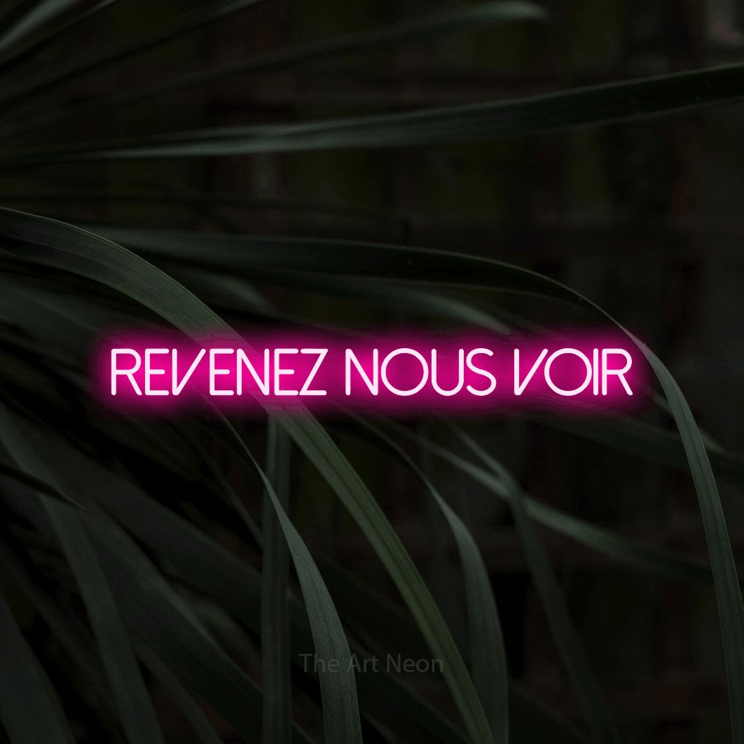 “Revenez nous voir” LED neon sign in hot pink, with thin sans-serif lettering, shown against a close-up of dark foliage.
