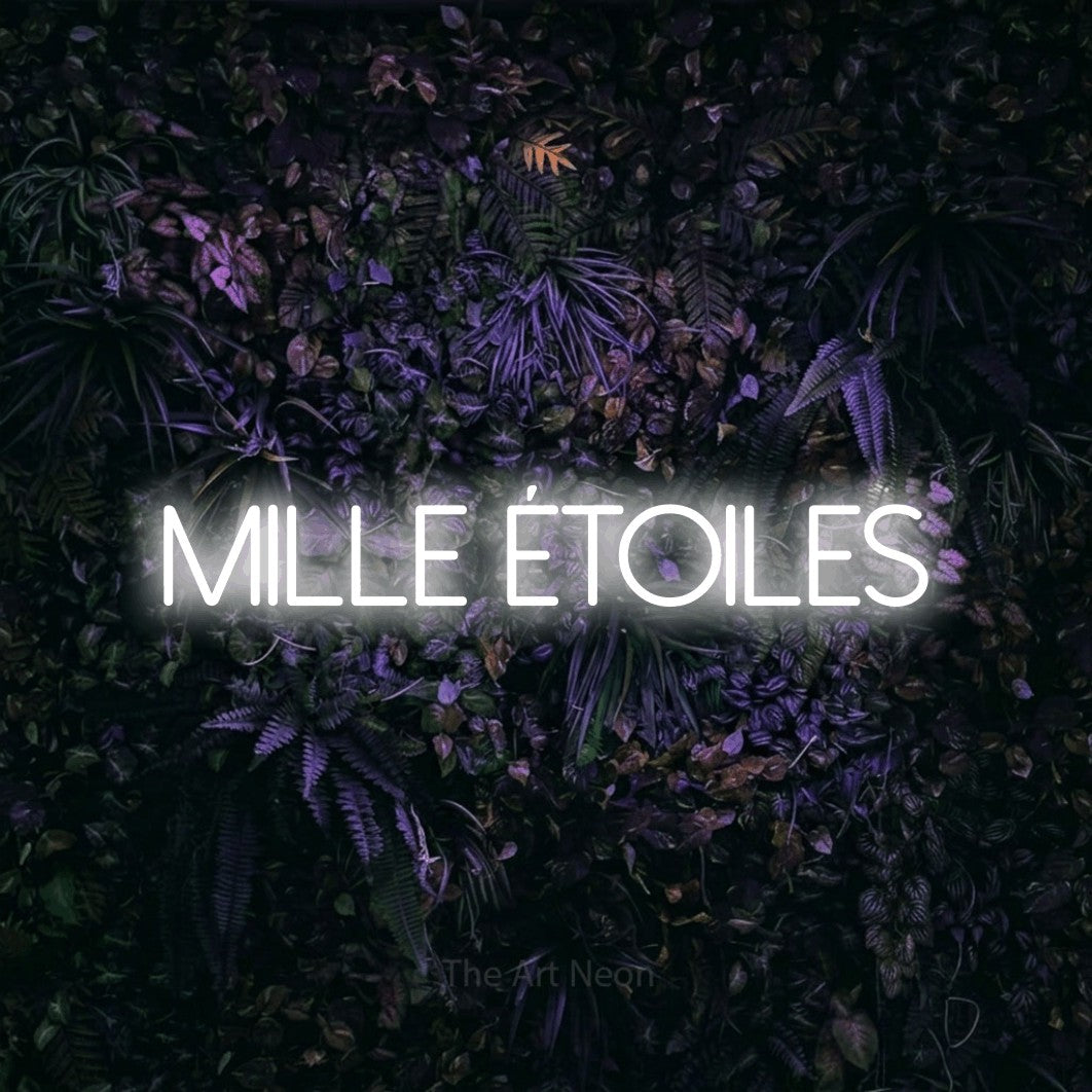 “Mille étoiles” LED neon sign in cool white, with thin sans-serif lettering, shown against a dark wall with purple flowers.