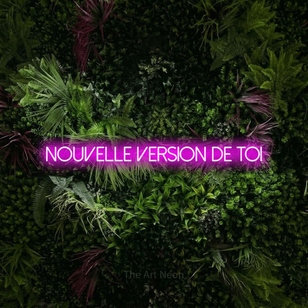 “Nouvelle version de toi” LED neon sign in pink, with thin sans-serif lettering, shown against a plant wall with pink flowers.