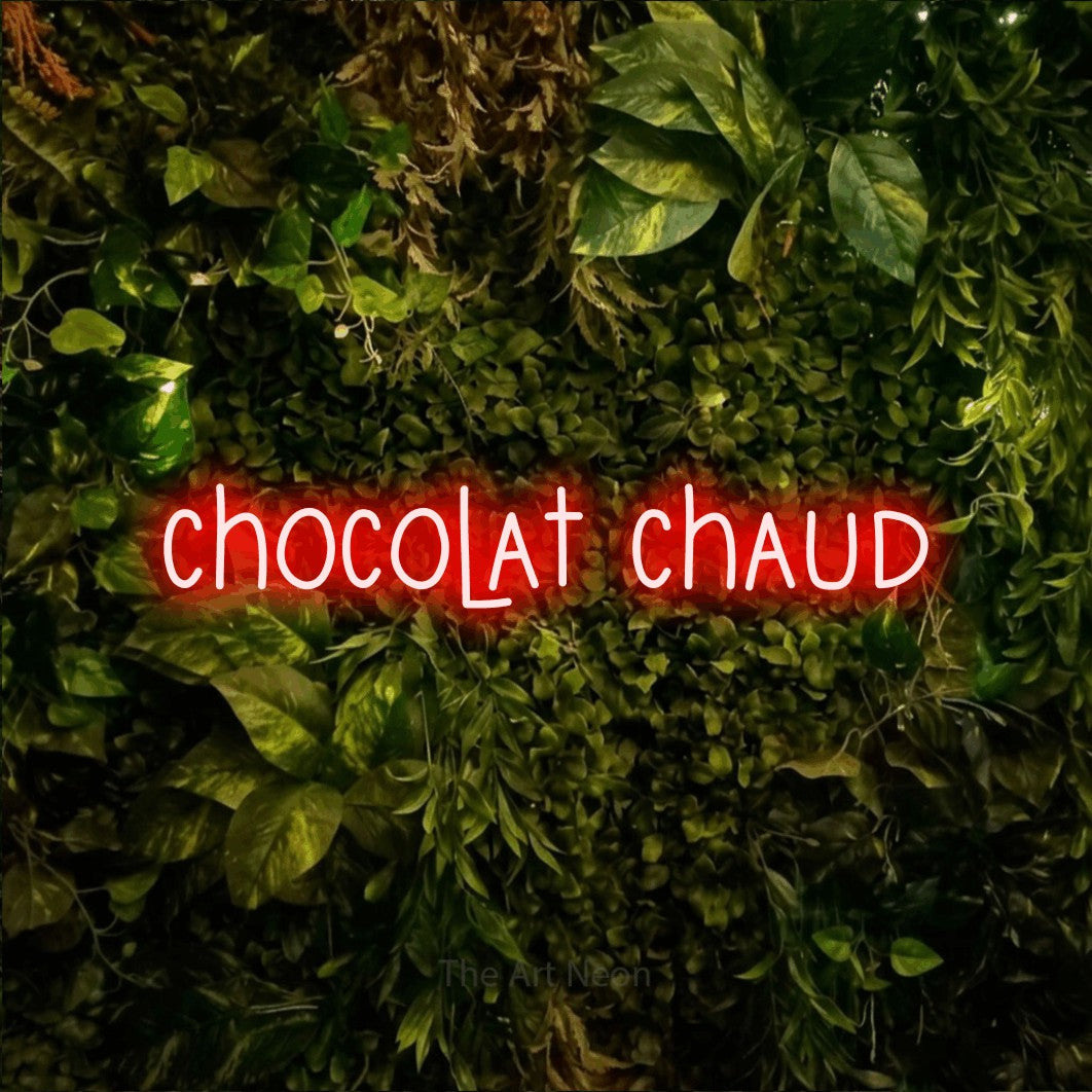 Décoration murale en néon LED « Chocolat chaud » en rouge, en lettres étroites dessinées à la main, installée sur un feuillage vert et jaune vif, pour un café.