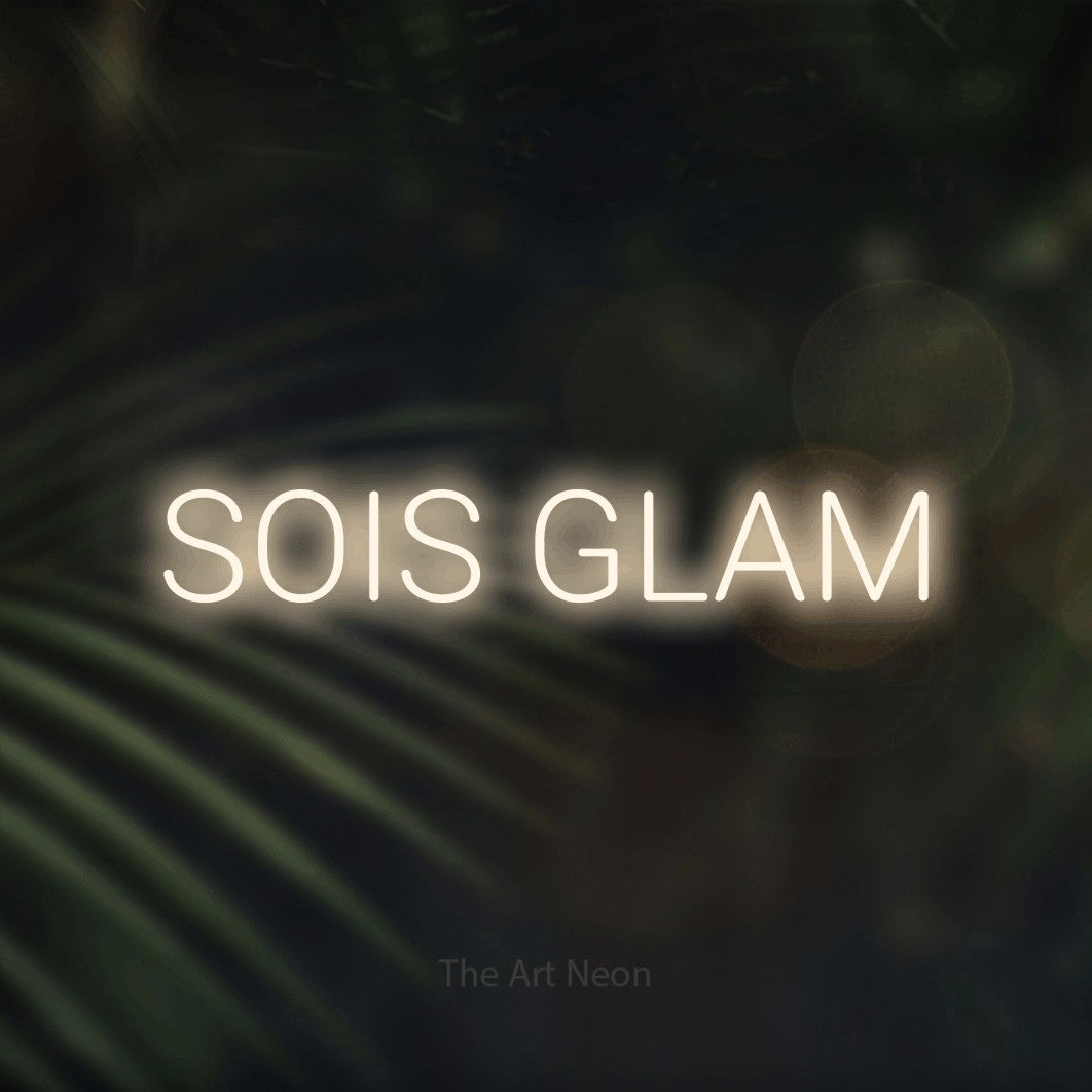 “Sois glam” LED neon sign in warm white, with rounded sans-serif lettering, shown against a blurred palm and bokeh-light backdrop.