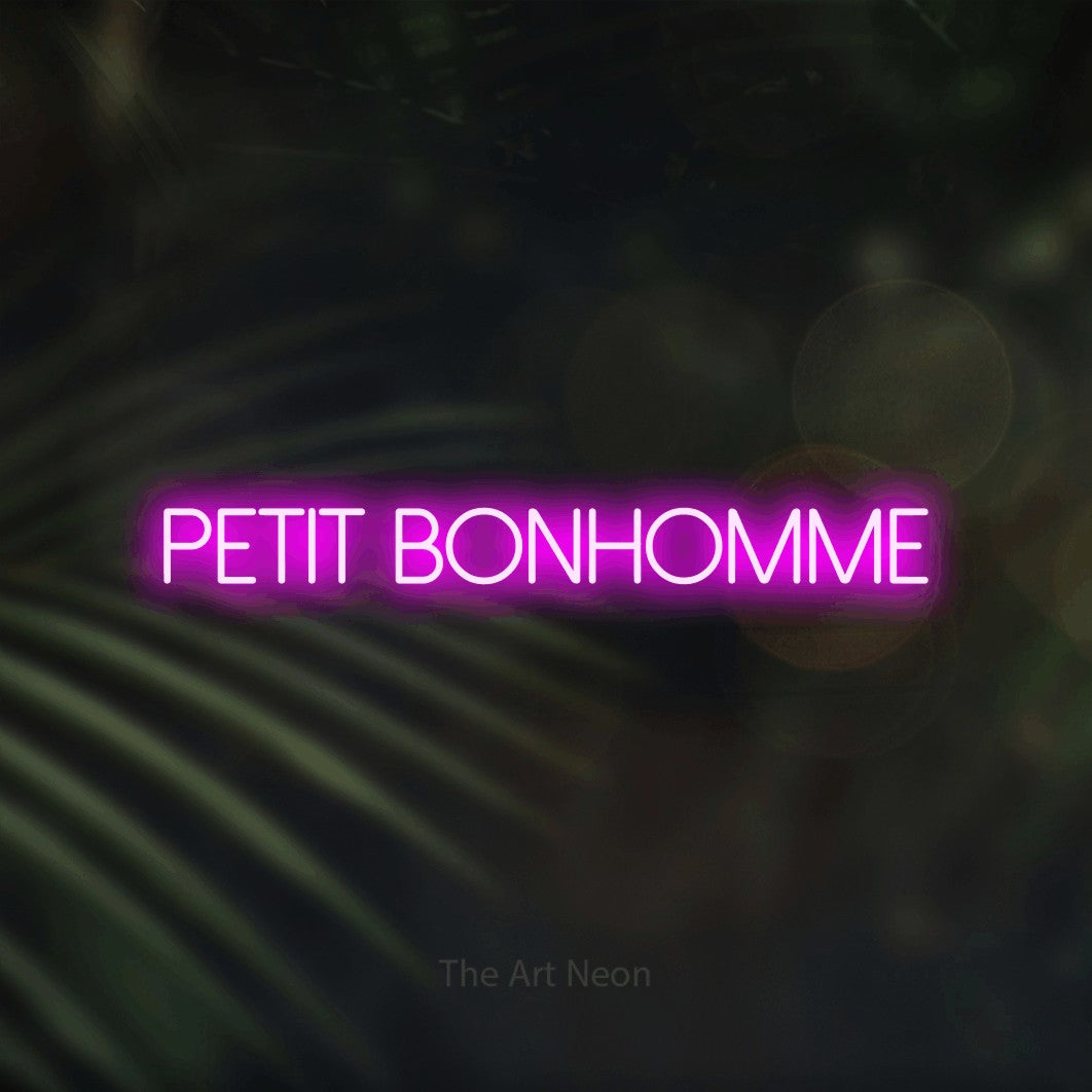 “Petit bonhomme” LED neon sign in pink, with thin sans-serif lettering, shown against a blurred palm and bokeh-light backdrop.