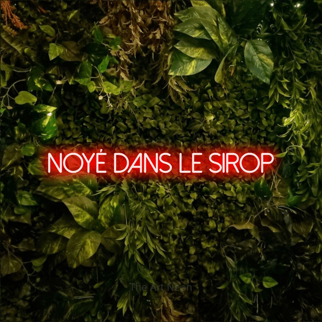 “Noyé dans le sirop” LED neon sign in red, with thin sans-serif lettering, shown against bright green and yellow foliage.
