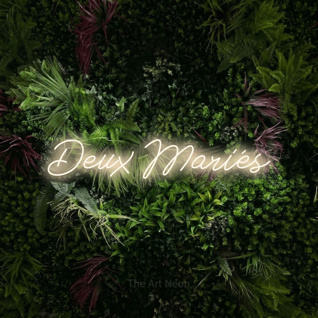 “Deux mariés” LED neon sign in warm white, with cursive lettering, shown against a plant wall with pink flowers.
