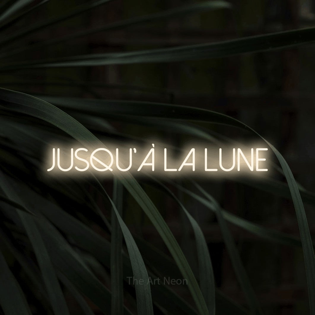 “Jusqu'à la lune” LED neon sign in warm white, with thin sans-serif lettering, shown against a close-up of dark foliage.