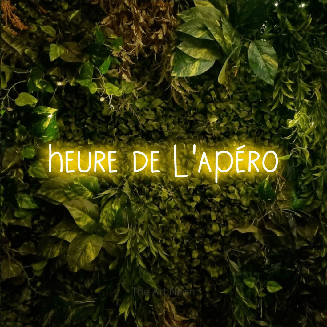 “Heure de l'apéro” LED neon sign in yellow, with narrow hand-drawn lettering, shown against bright green and yellow foliage.