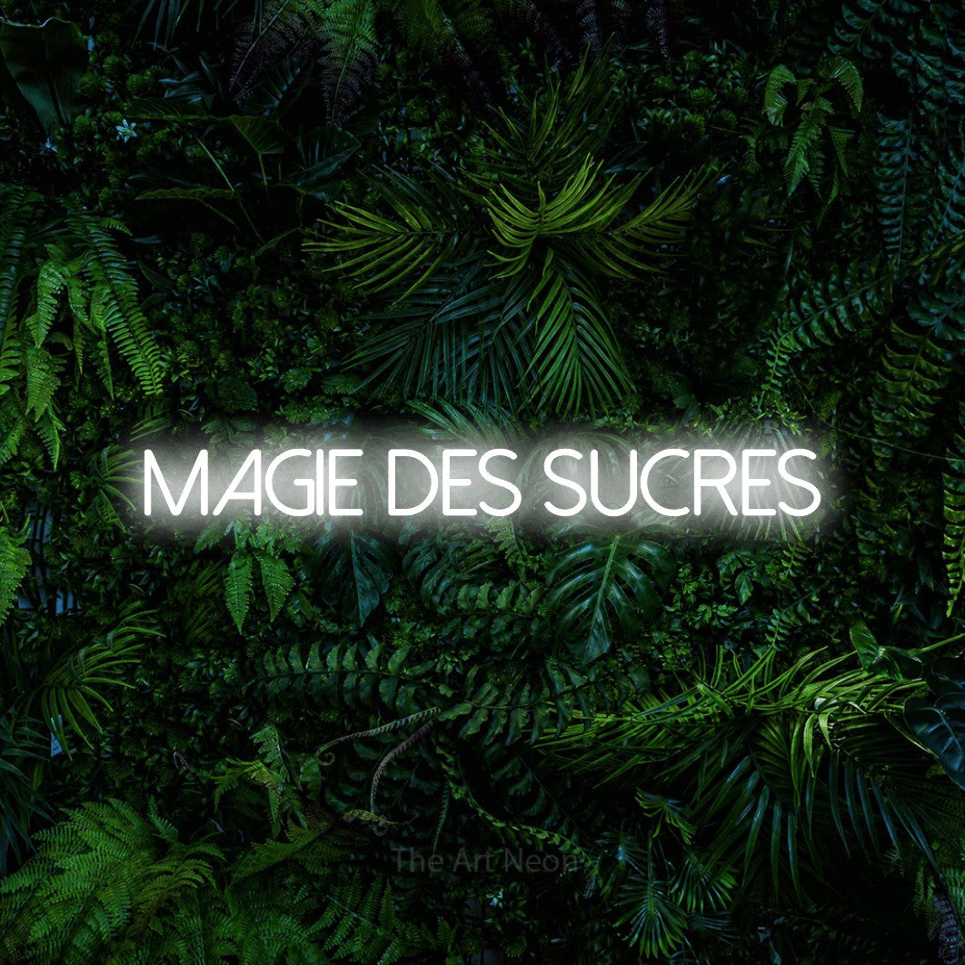 “Magie des sucres” LED neon sign in cool white, with thin sans-serif lettering, shown against dense tropical foliage.