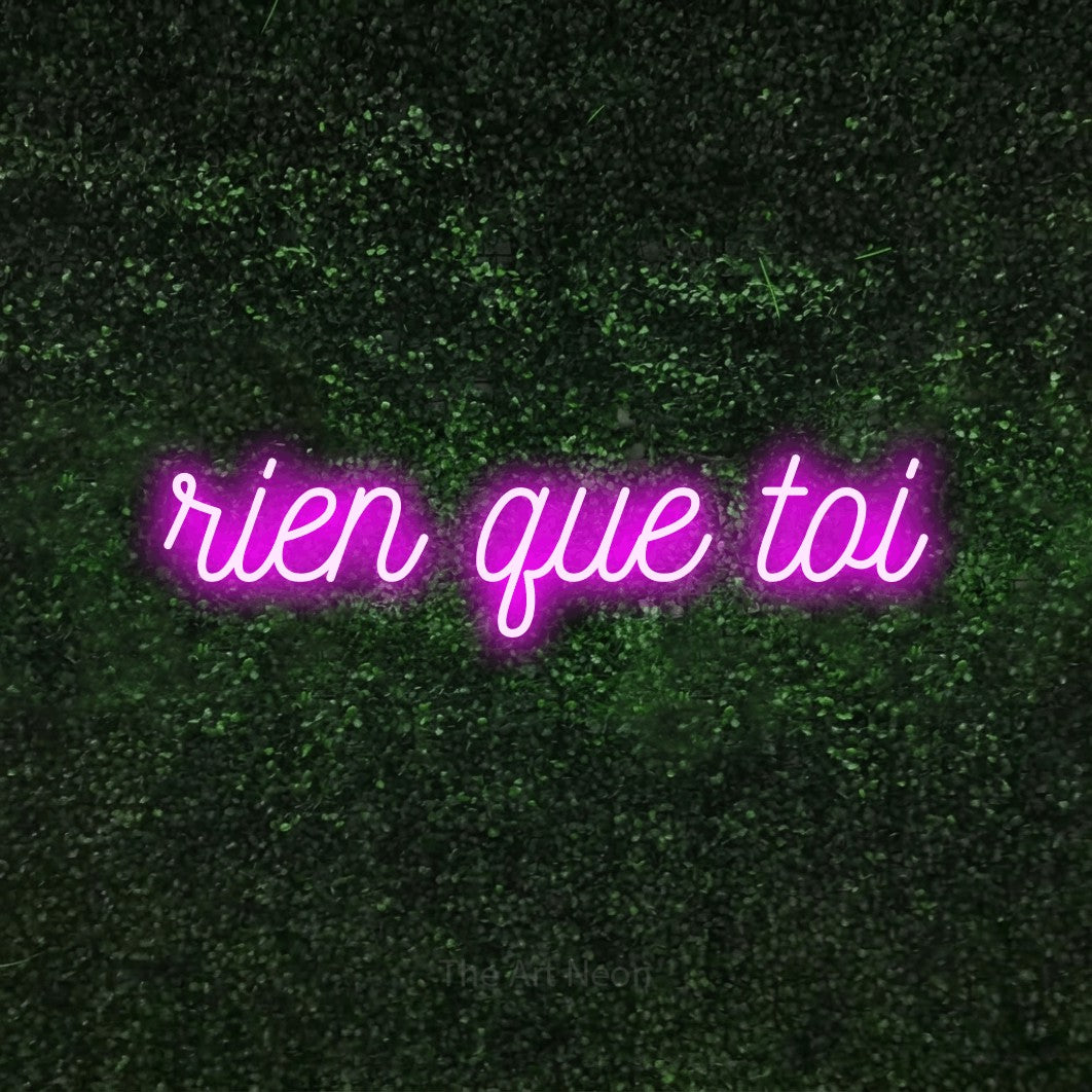 “Rien que toi” LED neon sign in pink, with ornate cursive lettering, shown against a textured green hedge.