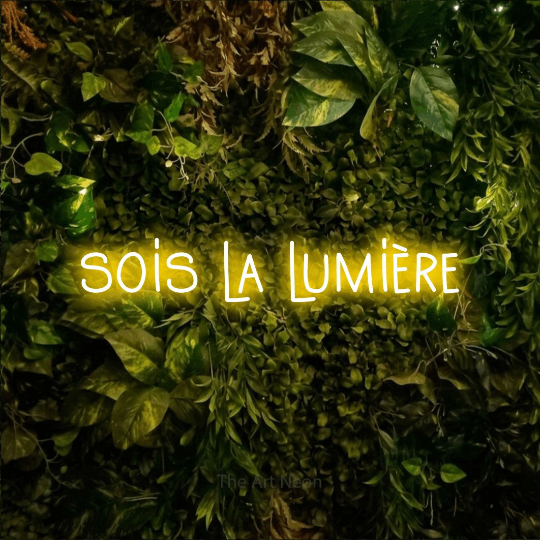 “Sois la lumière” LED neon sign in yellow, with narrow hand-drawn lettering, shown against bright green and yellow foliage.