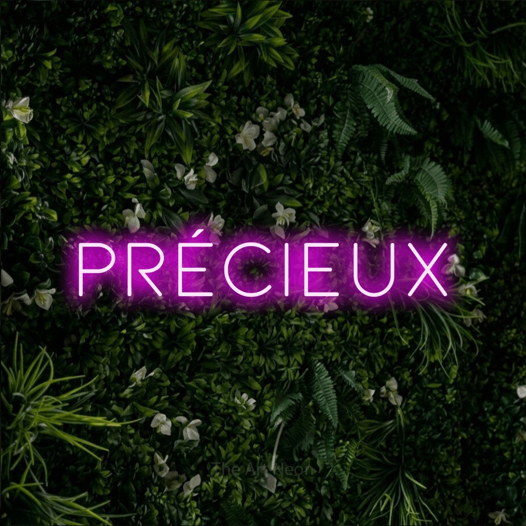 “Précieux” LED neon sign in pink, with thin, widely spaced lettering, shown against a dense green plant wall.
