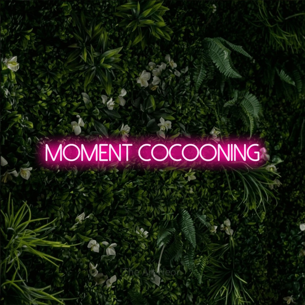 “Moment cocooning” LED neon sign in hot pink, with thin sans-serif lettering, shown against a dense green plant wall.