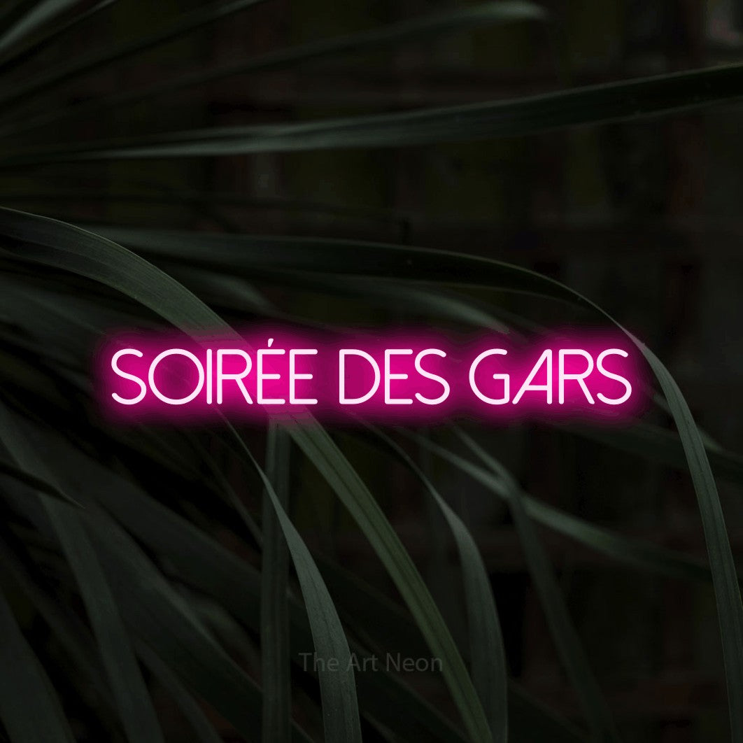 “Soirée des gars” LED neon sign in hot pink, with thin sans-serif lettering, shown against a close-up of dark foliage.