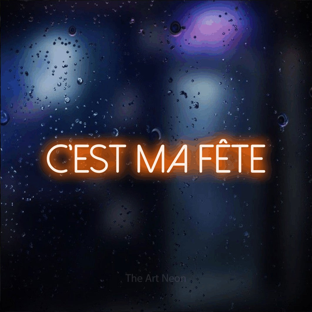 “C'est ma fête” LED neon sign in orange, with thin sans-serif lettering, shown against glass with blue and purple reflections.