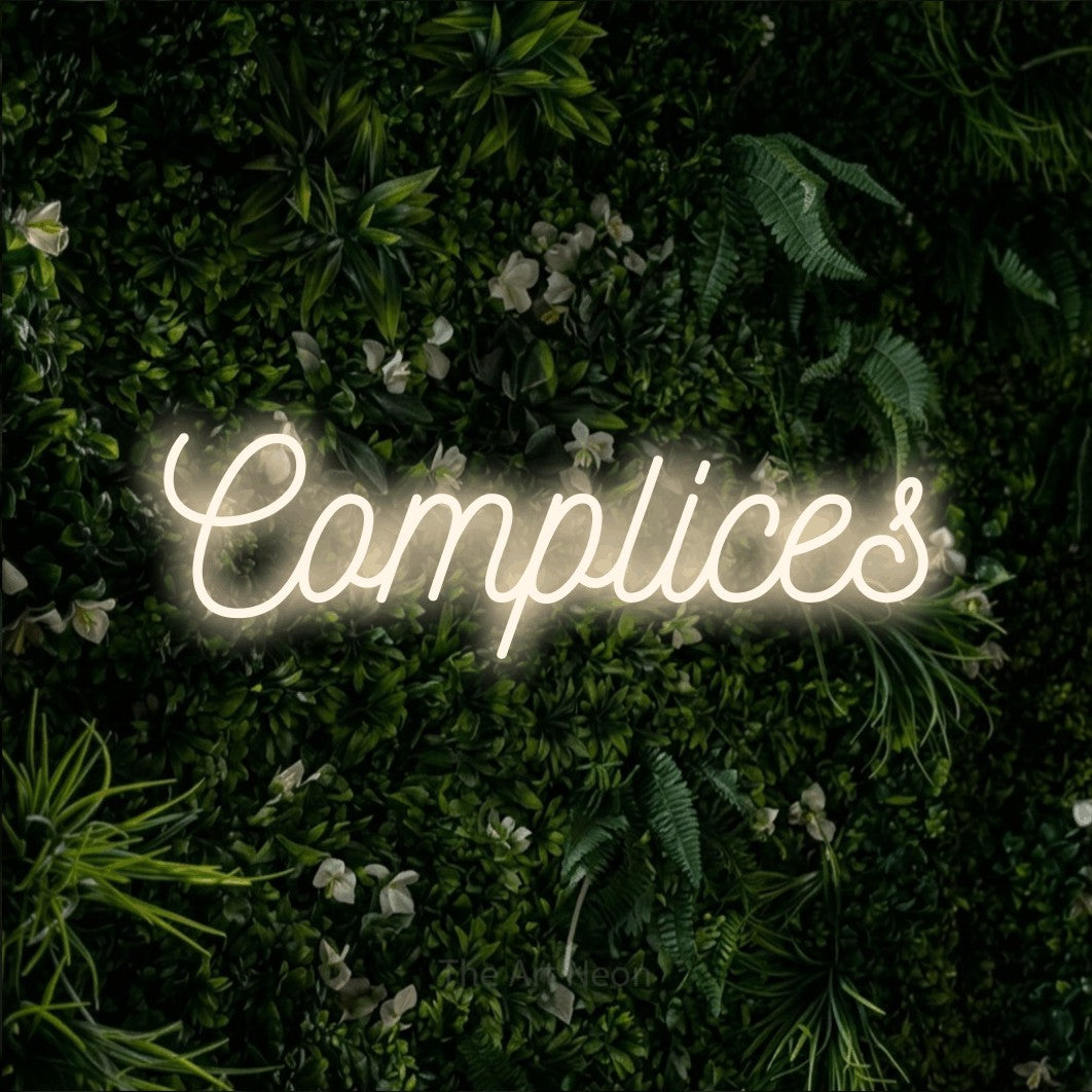 “Complices” LED neon sign in warm white, with ornate cursive lettering, shown against a dense green plant wall.