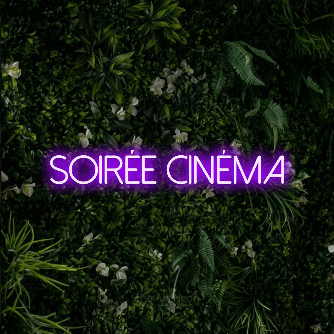 “Soirée cinéma” LED neon sign in purple, with thin sans-serif lettering, shown against a dense green plant wall.
