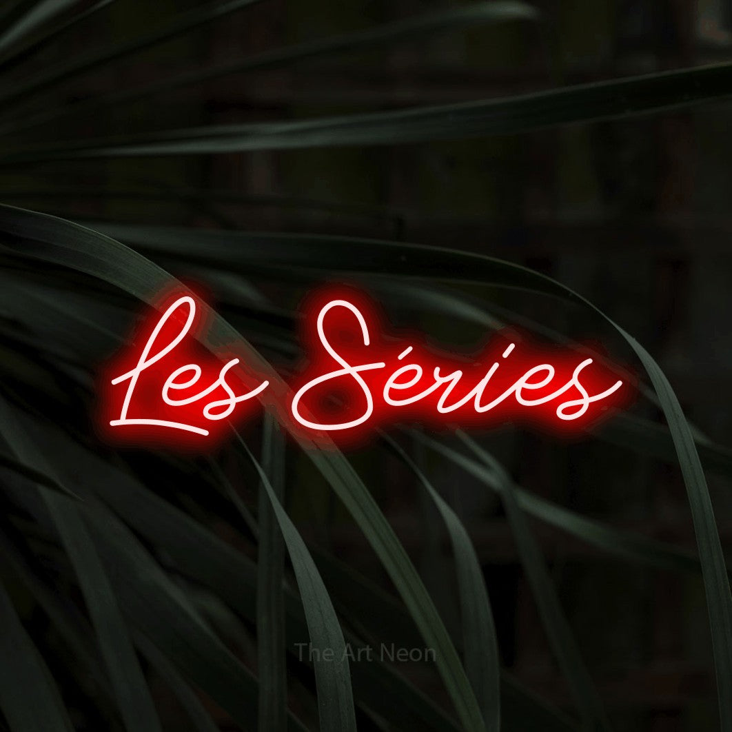 “Les séries” LED neon sign in red, with cursive lettering, shown against a close-up of dark foliage.