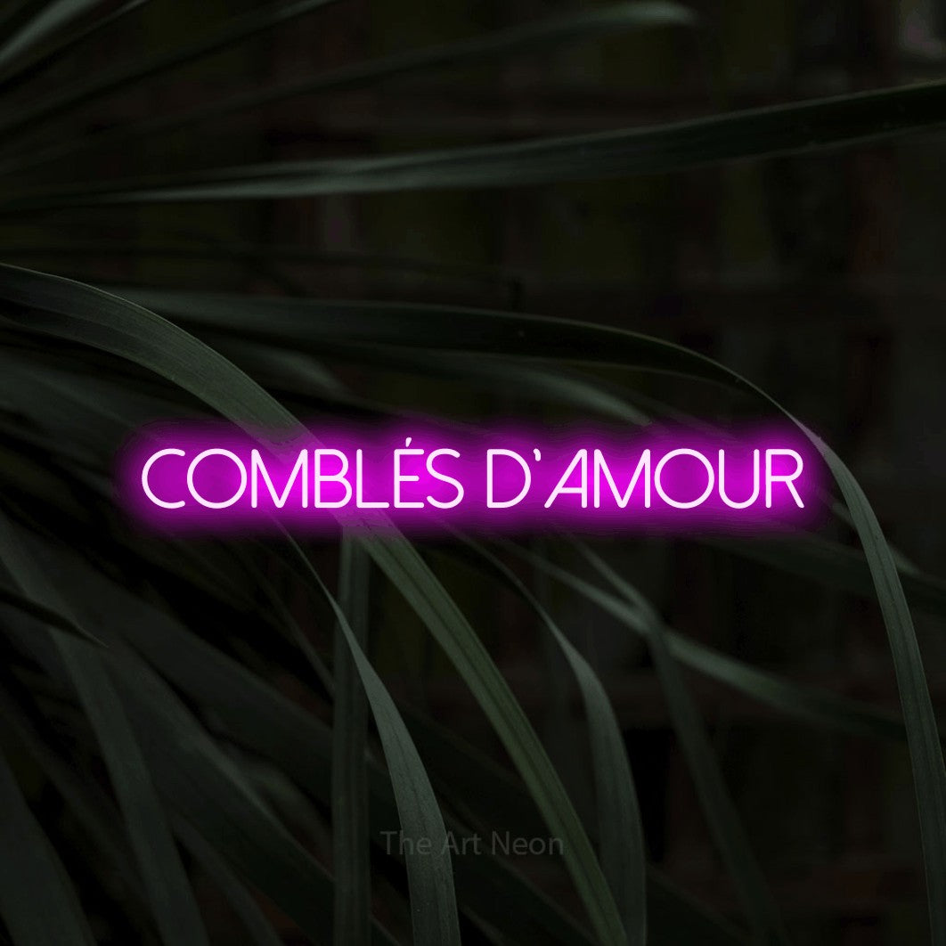 “Comblés d'amour” LED neon sign in pink, with thin sans-serif lettering, shown against a close-up of dark foliage.