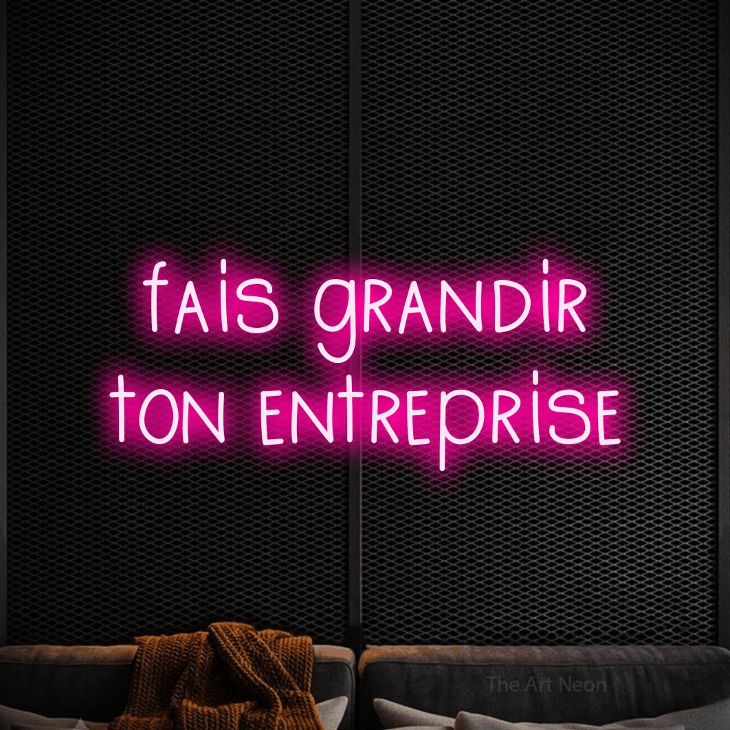 Enseigne néon LED « Fais grandir ton entreprise » sur deux lignes en rose vif, en lettres étroites dessinées à la main, présentée dans un coin salon moderne aux tons noirs, pour un bureau ou une entreprise.