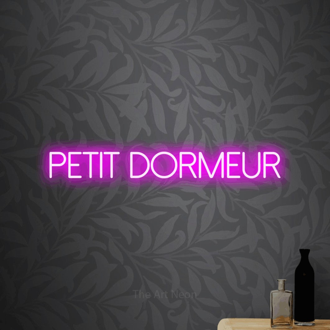 “Petit dormeur” LED neon sign in pink, with thin sans-serif lettering, shown against grey botanical wallpaper.