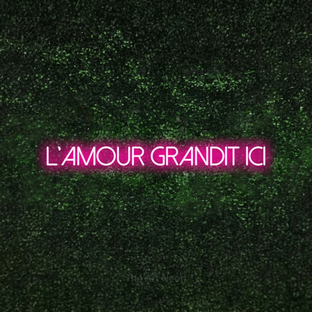 “L'amour grandit ici” LED neon sign in hot pink, with thin sans-serif lettering, shown against a textured green hedge.
