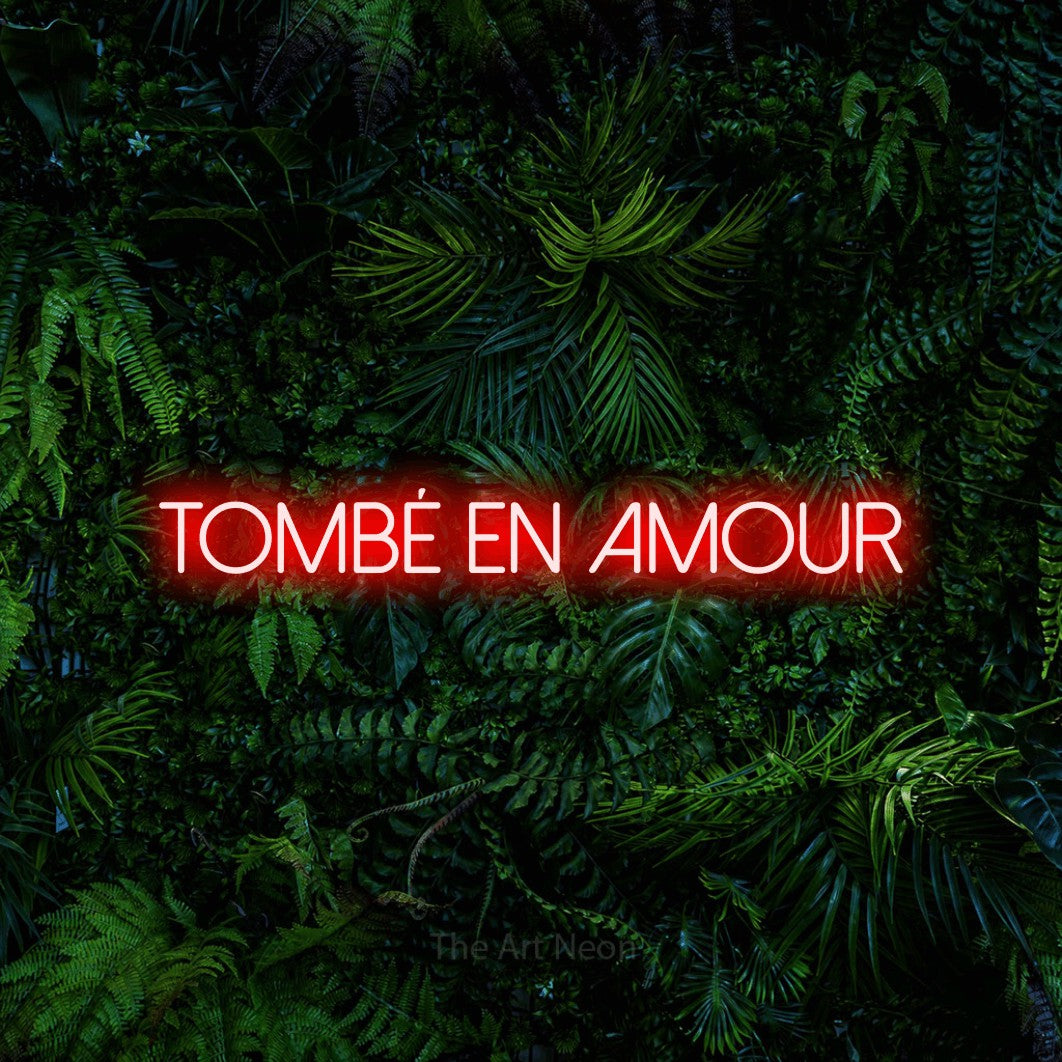 “Tombé en amour” LED neon sign in red, with thin sans-serif lettering, shown against dense tropical foliage.