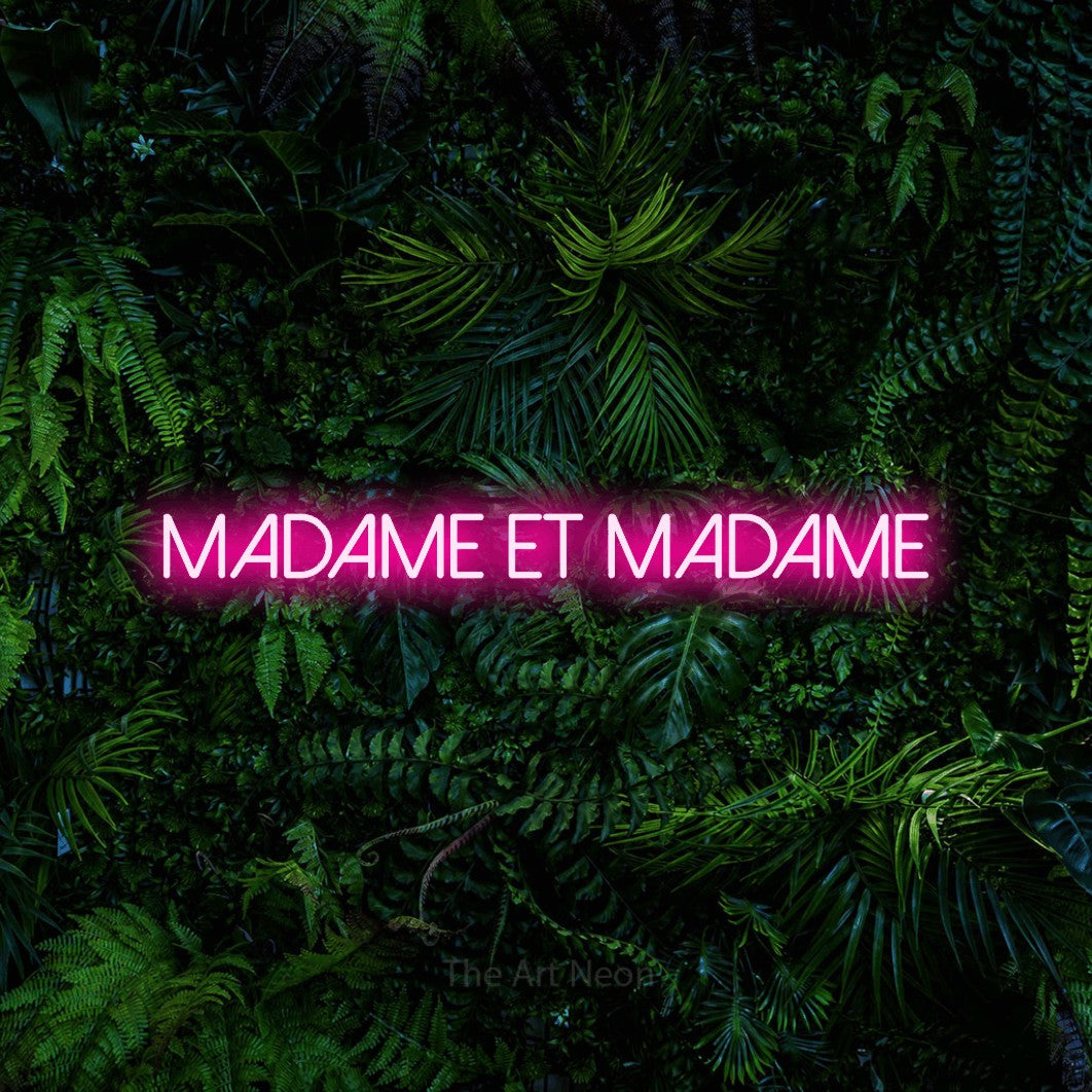 “Madame et madame” LED neon sign in hot pink, with thin sans-serif lettering, shown against dense tropical foliage.