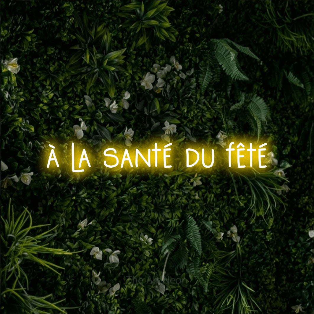 “À la santé du fêté” LED neon sign in yellow, with narrow hand-drawn lettering, shown against a dense green plant wall.