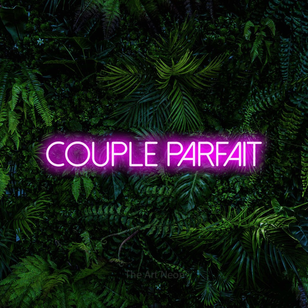 “Couple parfait” LED neon sign in pink, with thin sans-serif lettering, shown against dense tropical foliage.