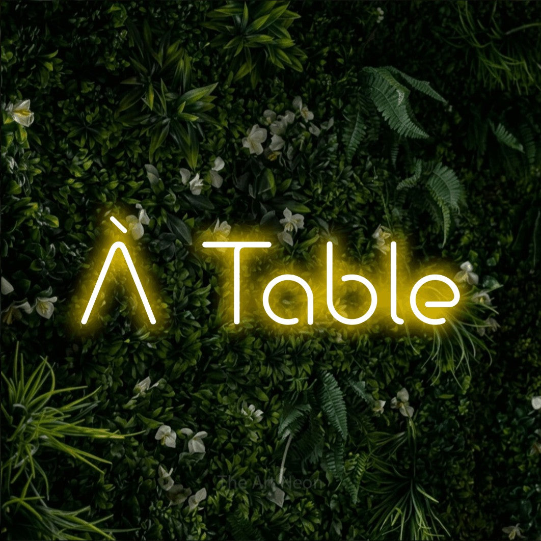 “À table” LED neon sign in yellow, with stylised geometric lettering, shown against a dense green plant wall.