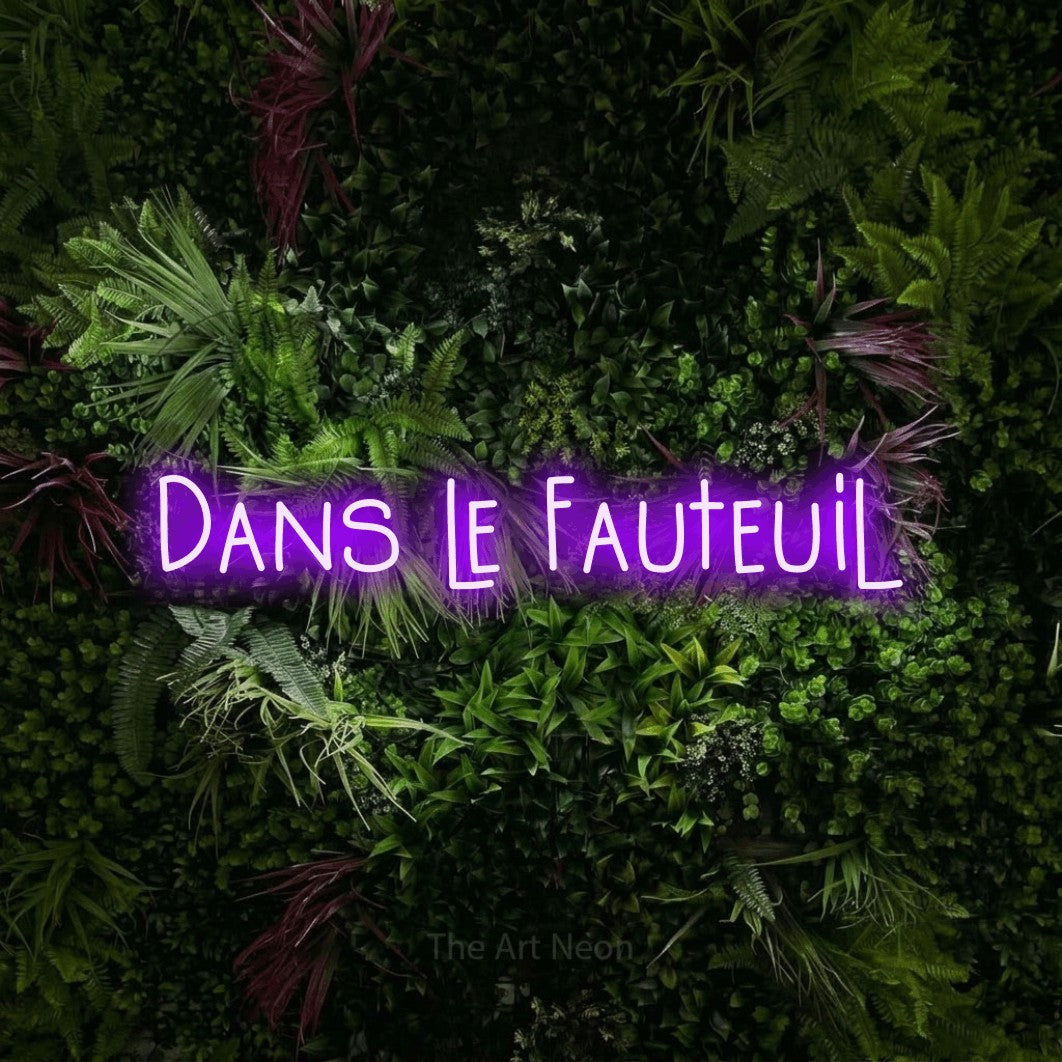 “Dans le fauteuil” LED neon sign in purple, with narrow hand-drawn lettering, shown against a plant wall with pink flowers.