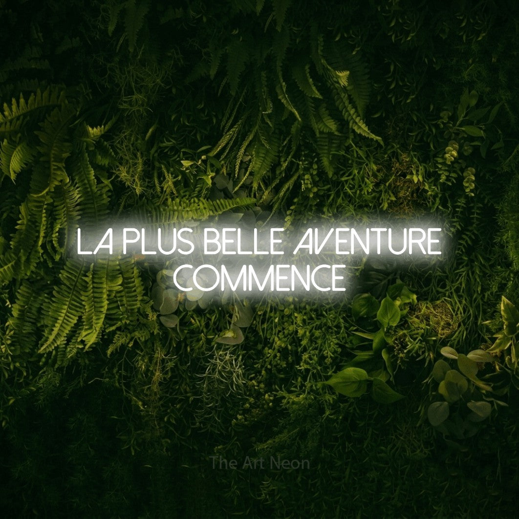 Composition en néon LED « La plus belle aventure commence » sur deux lignes en blanc froid, en lettres fines sans empattement, mise en valeur sur un fond sombre de mousse et de forêt, pour une fête prénatale.