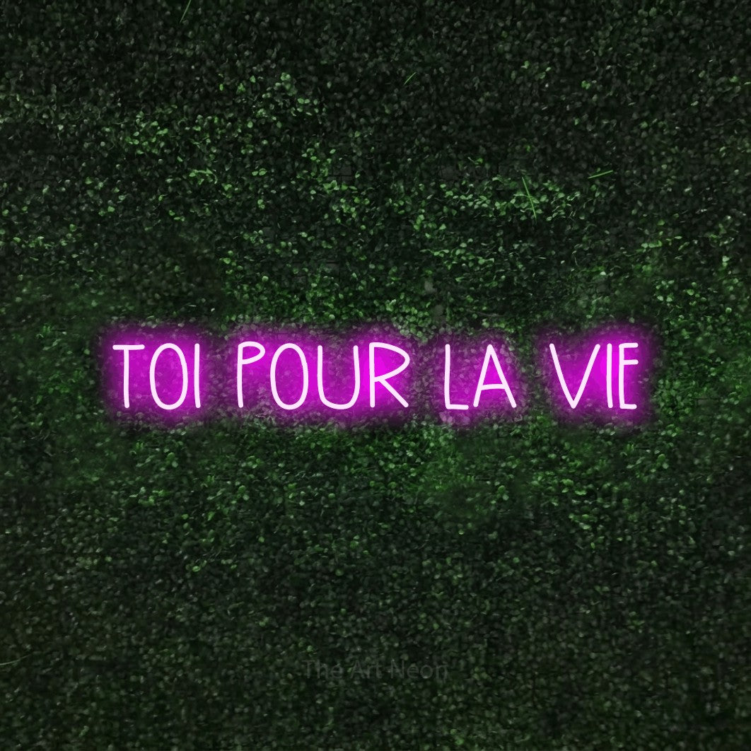 “Toi pour la vie” LED neon sign in pink, with narrow hand-drawn lettering, shown against a textured green hedge.