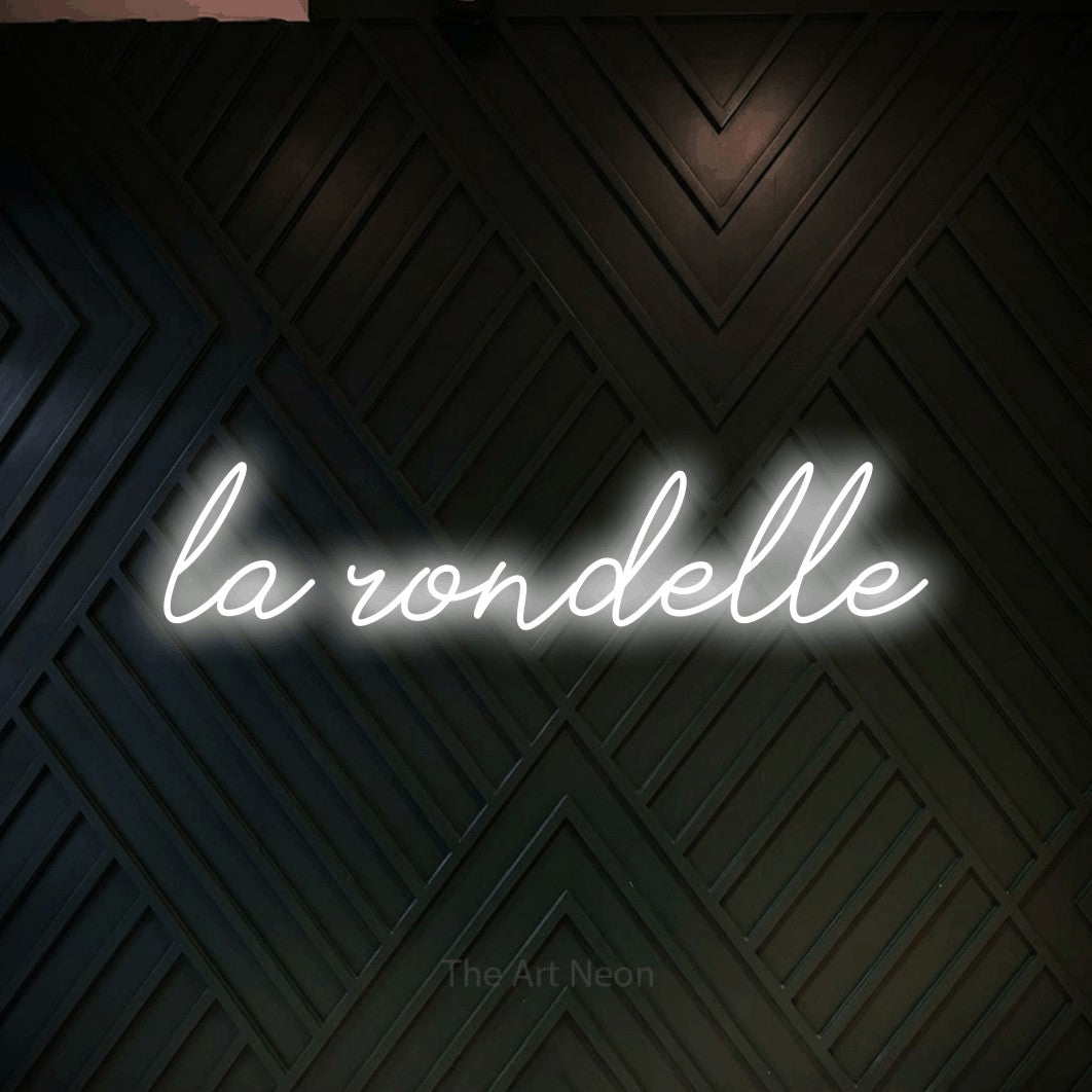 “La rondelle” LED neon sign in cool white, with cursive lettering, shown against a black chevron-patterned wood wall.