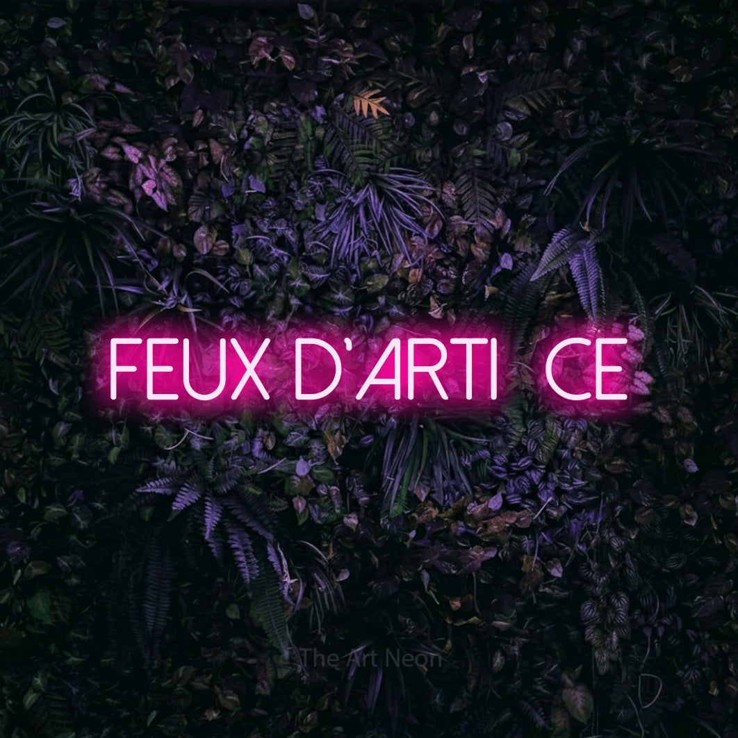 “Feux d'artifice” LED neon sign in hot pink, with thin sans-serif lettering, shown against a dark wall with purple flowers.