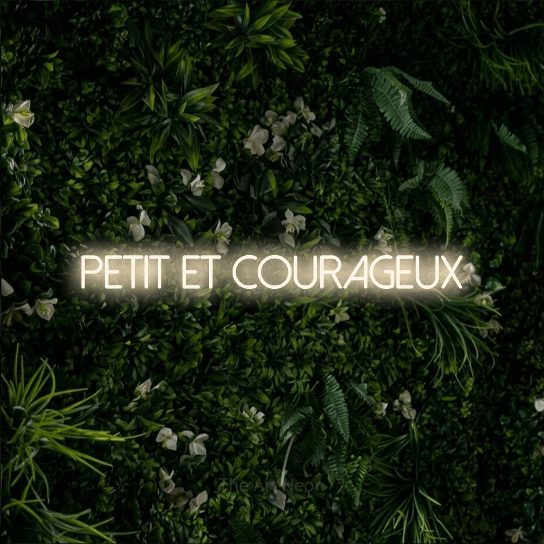 “Petit et courageux” LED neon sign in warm white, with thin sans-serif lettering, shown against a dense green plant wall.