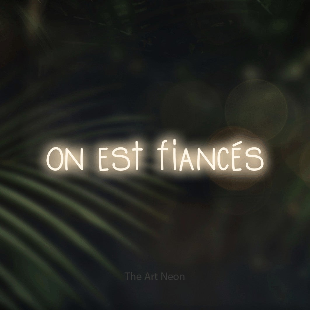 “On est fiancés” LED neon sign in warm white, with narrow hand-drawn lettering, shown against a blurred palm and bokeh-light backdrop.