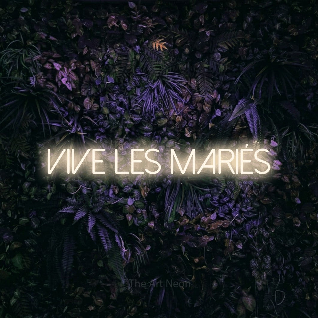 “Vive les mariés” LED neon sign in warm white, with thin sans-serif lettering, shown against a dark wall with purple flowers.