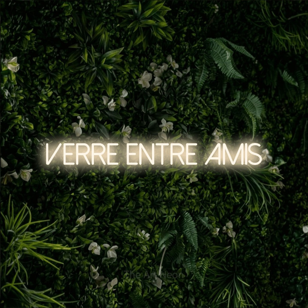 Composition en néon LED « Verre entre amis » en blanc chaud, en lettres fines sans empattement, mise en valeur sur un mur végétal vert et dense, pour un bar ou un pub.