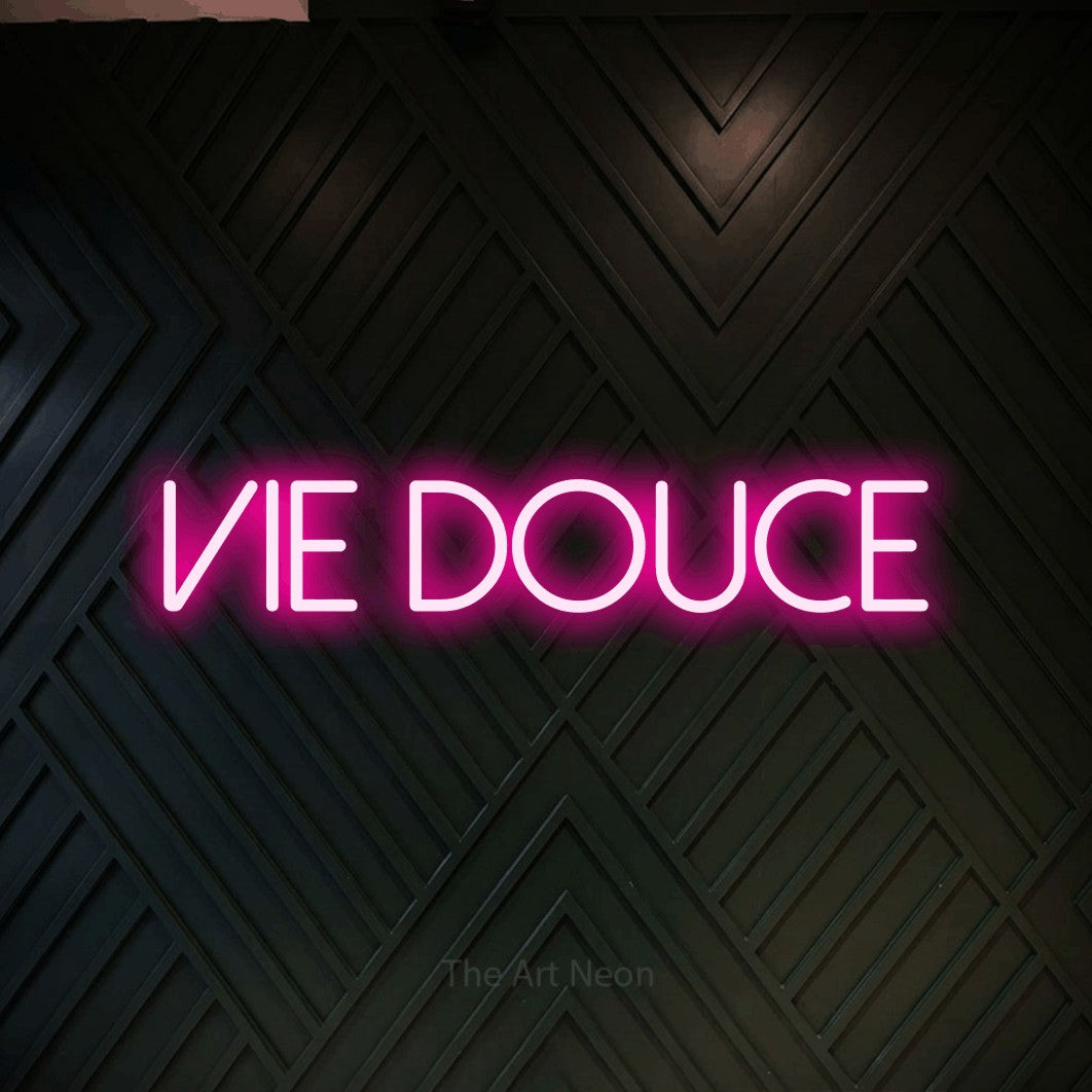 “Vie douce” LED neon sign in hot pink, with thin sans-serif lettering, shown against a black chevron-patterned wood wall.