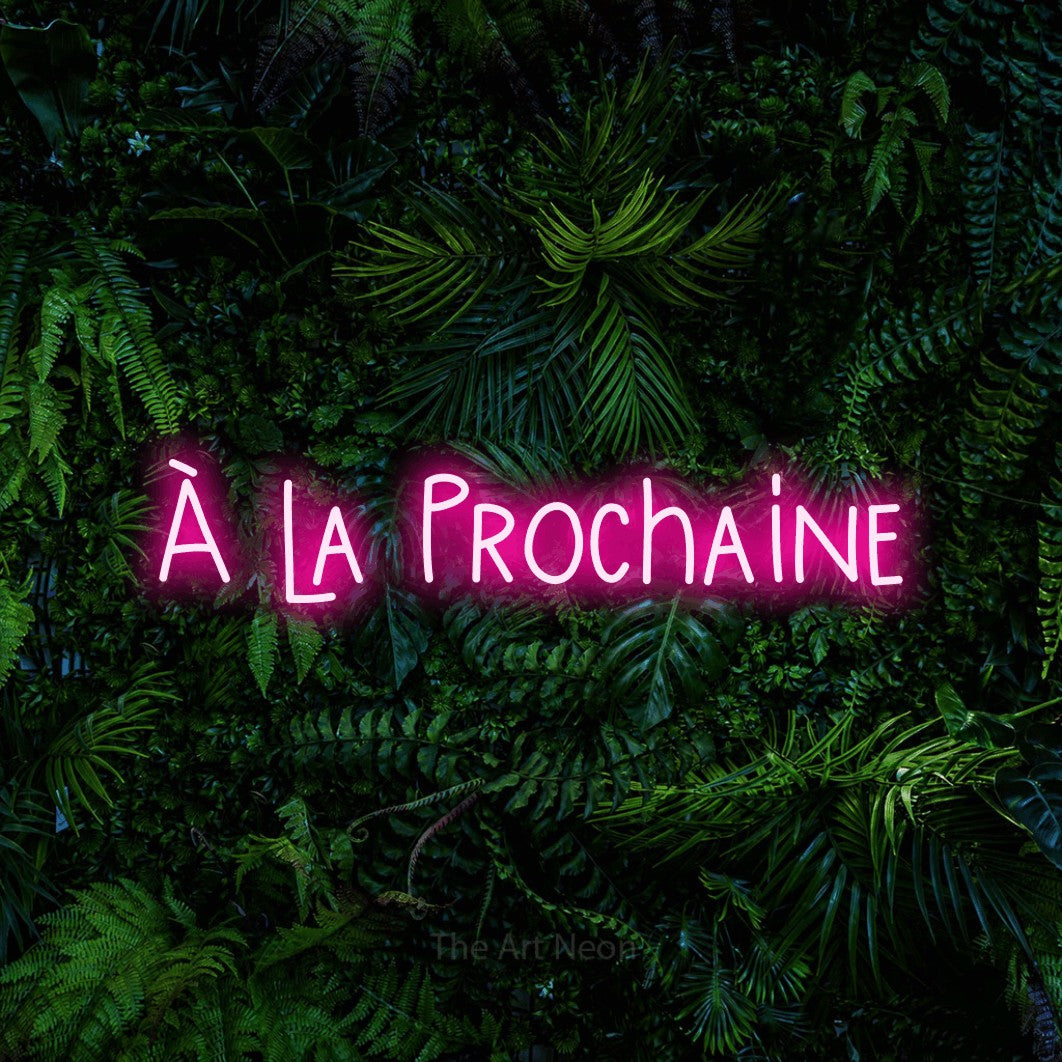“À la prochaine” LED neon sign in hot pink, with narrow hand-drawn lettering, shown against dense tropical foliage.